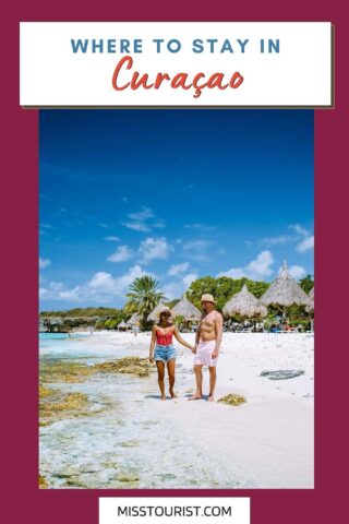A couple holding hands and walking along a sandy beach with clear blue water and thatched-roof structures in the background. The text reads, "WHERE TO STAY IN Curaçao.