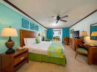 Bright turquoise guest room at Yellow Bird Hotel in Barbados, with a wooden bed, green and yellow bedding, ceiling fan and patio doors opening to a sea view.