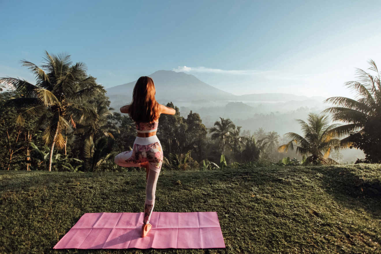 A woman doing yoga on a pink mat in a tropical landscape with palm trees and a mountain in the background.