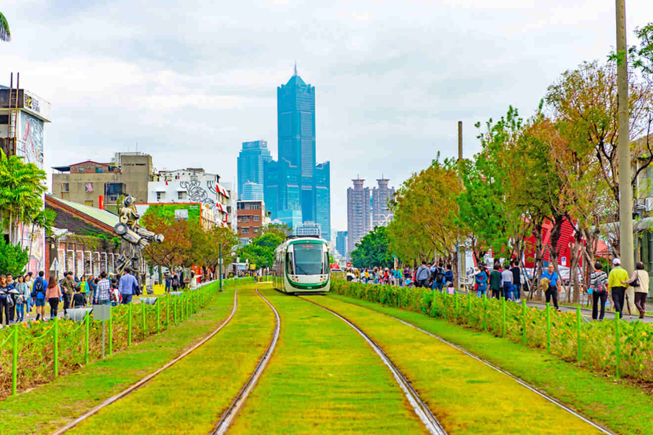 A modern tram travels on grassy tracks through an urban area with trees and buildings lining both sides, with a tall skyscraper visible in the background.
