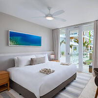 A minimalist bedroom with a large bed, white bedding, bedside tables with lamps, a ceiling fan, a landscape painting on the wall, and glass doors opening to a view of greenery and a building outside.