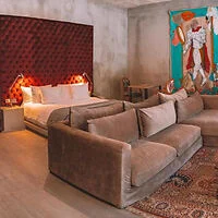 A hotel room featuring a king-sized bed with a red tufted headboard, a beige sectional sofa, a wall painting, and wooden furniture on light wood flooring with a patterned rug.