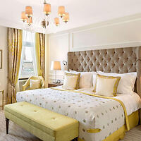 A bedroom with a large bed with a tufted headboard, yellow bedding, and pillows. A bench is at the foot of the bed. There are yellow curtains, a window, and a chandelier above.