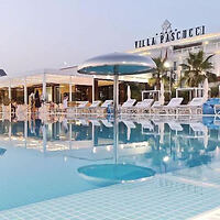 Outdoor swimming pool at Villa Pascucci with lounge chairs and umbrellas surrounding the pool. The building and name sign of Villa Pascucci are visible in the background.