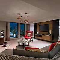 A modern living room with a red armchair, grey sectional sofa, and a round coffee table. A large television is mounted on a wooden wall unit. Floor-to-ceiling windows showcase a nighttime city view.