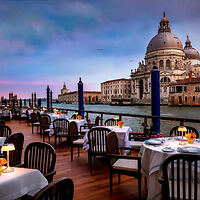 Outdoor restaurant with tables set along a waterfront, overlooking a historic domed building and a canal at dusk.
