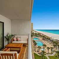 Balcony view of a beach resort featuring a seating area with a wooden table and chairs, overlooking pools, palm trees, and a sandy beach with clear blue skies and ocean in the background.