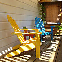 A small deck area with yellow and blue Adirondack chairs placed side by side, casting shadows from the slatted roof above. ​​