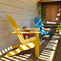 A small deck area with yellow and blue Adirondack chairs placed side by side, casting shadows from the slatted roof above. 