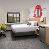 Modern hotel room with a king-sized bed, a wall-mounted lifebuoy decoration, a window with a view, a desk, a green chair, and a small refrigerator.