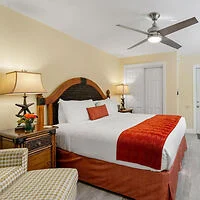Hotel room with a large bed featuring white linens and an orange throw blanket. The room includes two lamps on bedside tables, a ceiling fan, and a checkered armchair. Walls are painted light yellow.