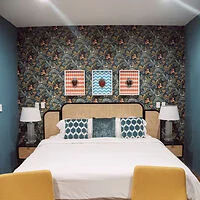 A bedroom with a double bed, two bedside tables with lamps, a floral-patterned wallpaper, and three framed pieces of art above the headboard. The bed has white sheets and teal pillows.