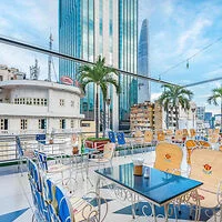 A rooftop patio with chairs and tables overlooks a cityscape with modern high-rise buildings and palm trees under a partly cloudy sky.