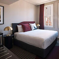 A modern bedroom featuring a neatly made bed with white sheets and reddish-brown pillows, flanked by two side tables with brass lamps. Two windows with dark curtains allow in natural light.