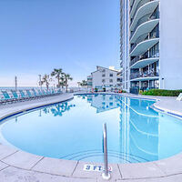 Outdoor swimming pool next to a multi-story building with balconies, surrounded by lounge chairs. Ocean and a clear sky in the background.