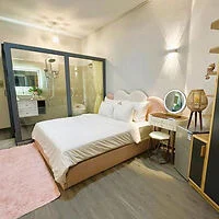 A modern bedroom with a double bed, a glass-enclosed bathroom, a vanity table with a round mirror, ambient lighting, and a pink area rug.
