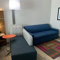 A small seating area with a blue sofa, a chair, a blue ottoman, and a small round orange table. A floor lamp and a piece of framed artwork are on the wall behind the sofa.