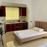 A small, tidy room with a single bed, a desk, and a kitchenette with red cabinets. A window with a yellow blind is above the sink area.