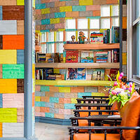 A colorful reading nook with a wall of multi-colored bricks, bookshelves filled with books and toys, and two black chairs with orange cushions. A bouquet of flowers is placed near the chairs.