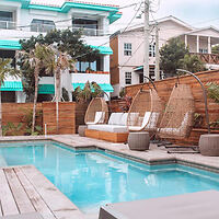 A modern outdoor pool area with hanging chairs, a cushioned bench, and surrounding wooden deck, set against the backdrop of a multi-story house.