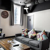 A modern living room features a gray sofa with colorful pillows, a wooden coffee table, a wall clock, and an industrial-style ceiling vent.