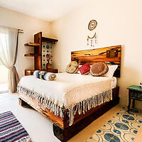 A cozy bedroom featuring a wooden bed with pillows, a throw, and a blanket. The room has a small shelf, a side table, patterned rug, and large window with curtains.