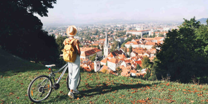 A person with a hat and yellow backpack stands beside a bicycle, overlooking a city with red-roofed buildings from a grassy hilltop.