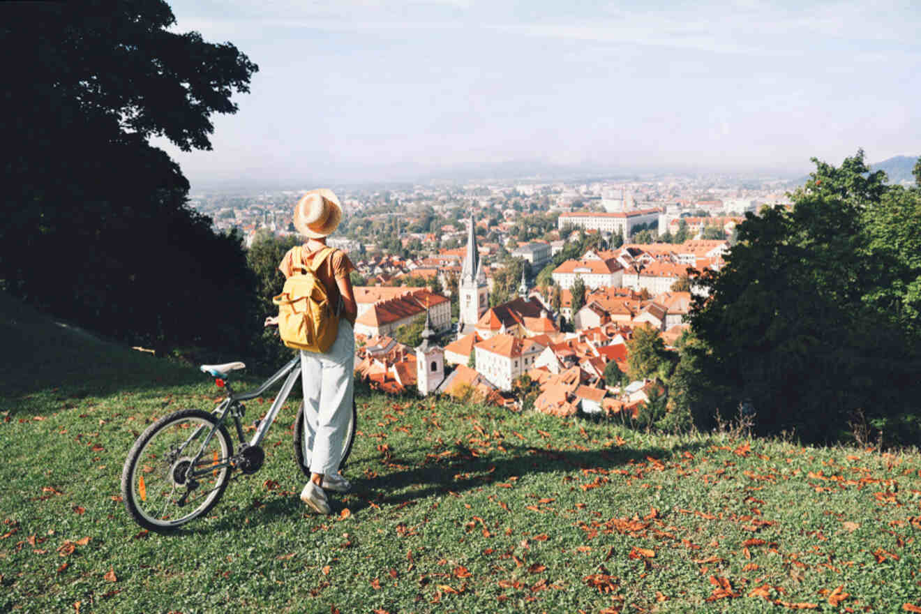 A person with a hat and yellow backpack stands beside a bicycle, overlooking a city with red-roofed buildings from a grassy hilltop.