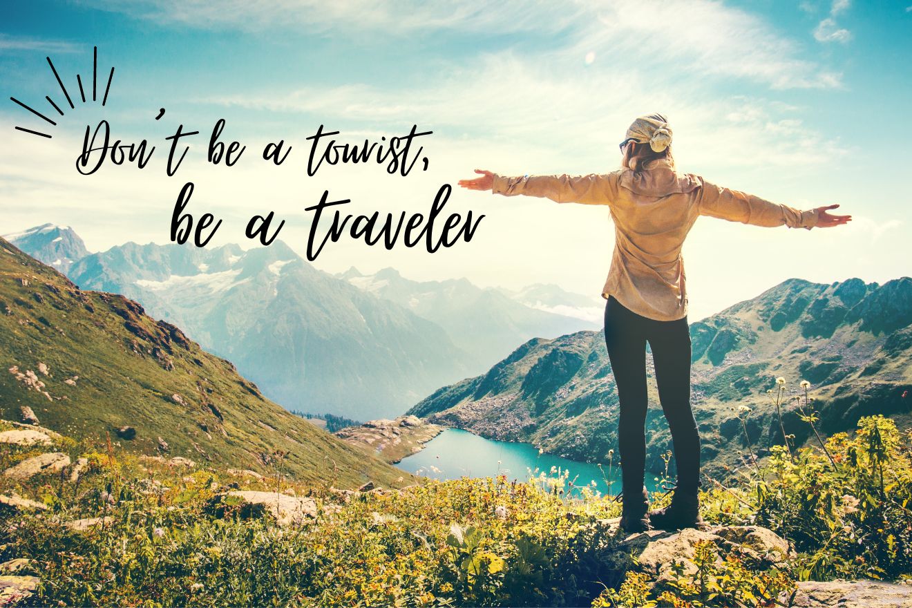 A person stands with arms outstretched on a grassy mountain overlooking a valley and lake, with the text "Don't be a tourist, be a traveler.