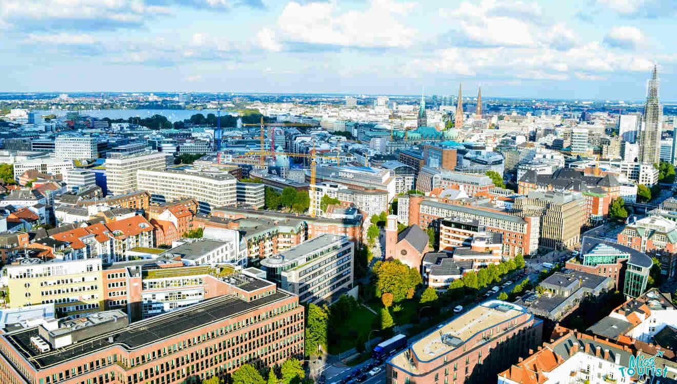 Aerial view of a cityscape featuring a mix of residential and commercial buildings, tree-lined streets, and distant water bodies under a partly cloudy sky.