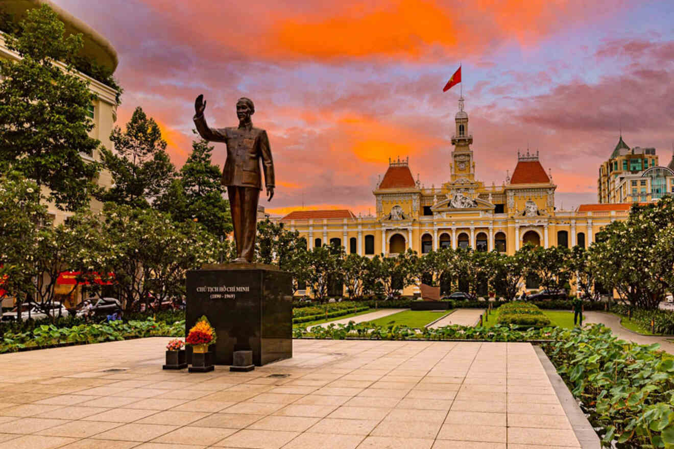 Statue of a man in front of a historical building with a red flag on top, surrounded by trees, under a vibrant orange sunset sky.
