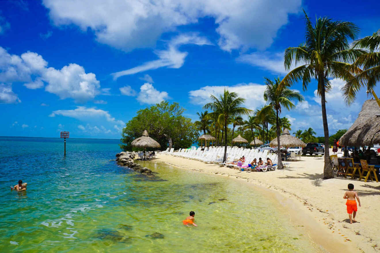 A tropical beach scene with clear blue water, palm trees, thatched umbrellas, white lounge chairs, and people swimming, sunbathing, and relaxing.