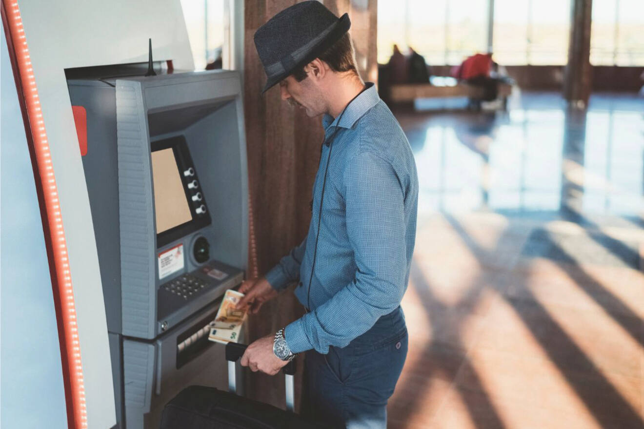 A man wearing a hat and blue shirt withdraws cash from an ATM inside a well-lit modern building, holding banknotes in his hand.