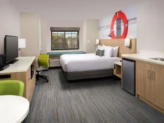 A hotel room with a queen bed, green chair, desk, TV, mini fridge, wall art featuring a lifebuoy, and large window with a built-in bench.