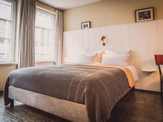 A neatly made bed with a gray blanket and white pillows in a cozy bedroom with large windows, light-colored walls, and framed artwork above the headboard.