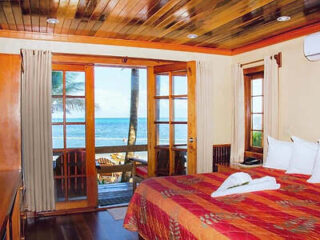 A cozy bedroom with a striped bedspread, wooden ceiling, and doors opening to a deck with an ocean view.