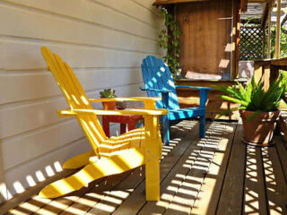 wo colorful Adirondack chairs, one yellow and one blue, on a sunny wooden porch with potted plants and wooden decor.