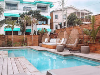 Outdoor pool area with wooden decking, lounge chairs, wicker hanging chairs, and adjacent palm trees. Two multi-story buildings are in the background, along with a wooden privacy fence.