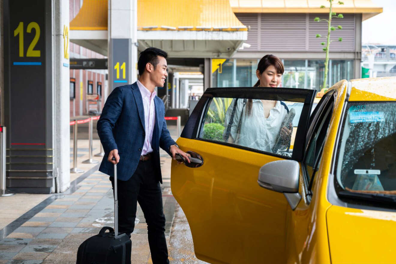 A man in a suit and carrying a suitcase helps a woman into a yellow taxi at an airport terminal, with numbered pillars in the background.