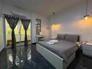 A modern bedroom with a double bed, dark bedding, two side tables, hanging lights, a small desk, and a balcony with a sliding glass door, all set on a glossy dark-tiled floor.