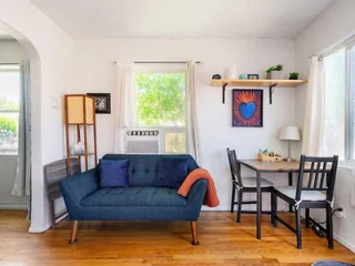 A small living room with a blue sofa, a table with two chairs, and a shelf with decor items. There is a window with curtains and an air conditioner. The wooden floor adds warmth to the space.