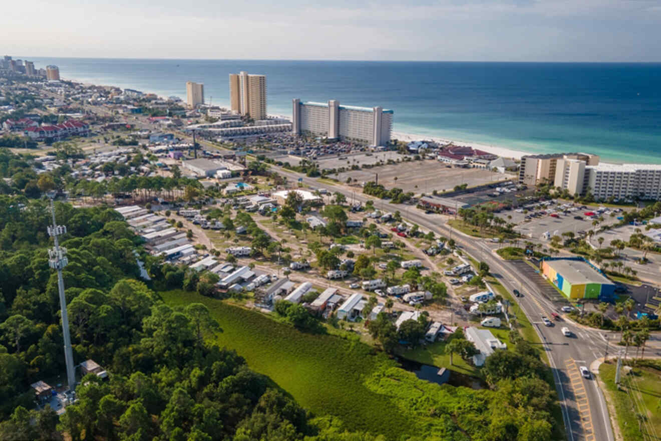 Aerial view of a coastal city with high-rise buildings near the beach, numerous parked vehicles, and lush green areas. The beach is visible along with blue ocean water.