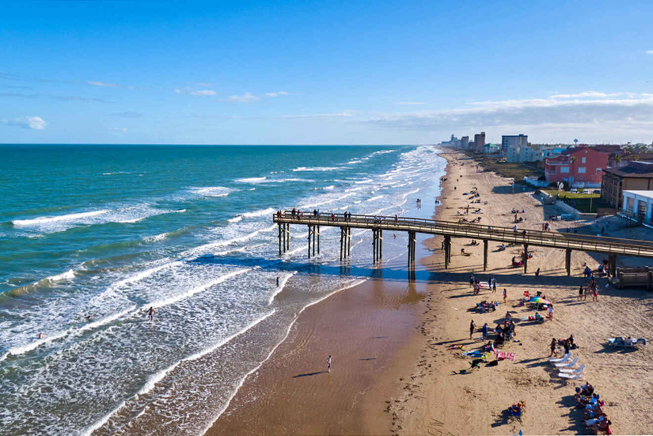 Aerial view of a beach with a pier extending into the ocean. People are on the sand and in the water. Buildings line the coastline in the background under a clear blue sky.