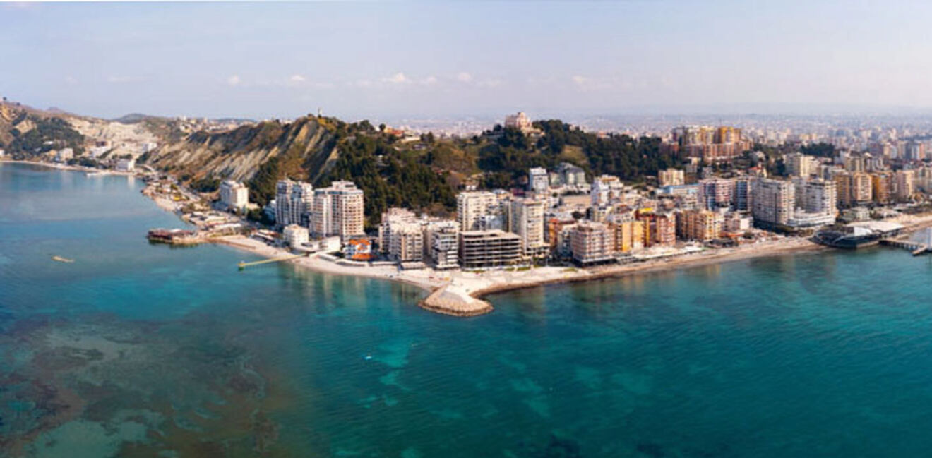 A coastal city with numerous buildings bordered by the sea, clear waters, and hilly terrain in the background under a clear sky.