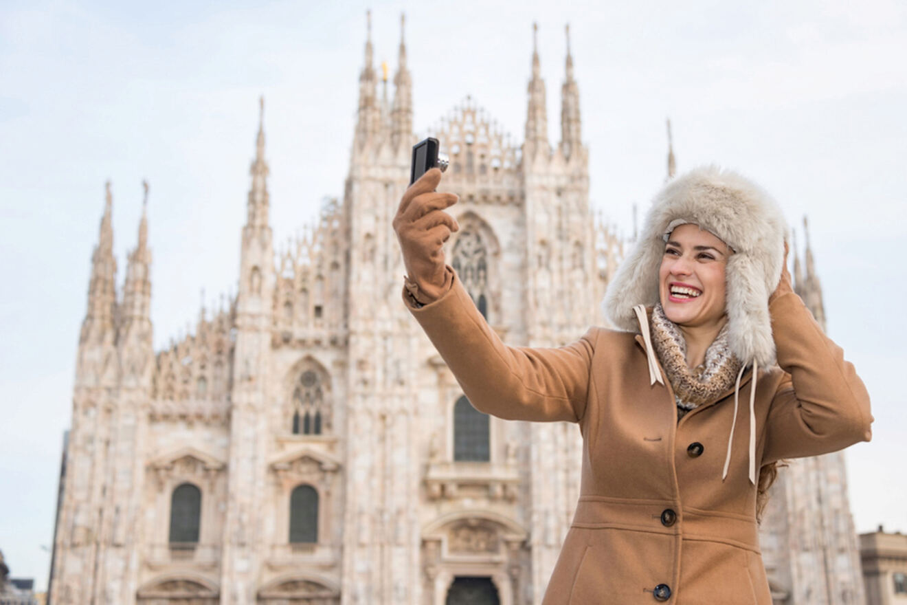 A woman in a tan coat and fur hat takes a selfie in front of a large, ornate cathedral with pointed spires.