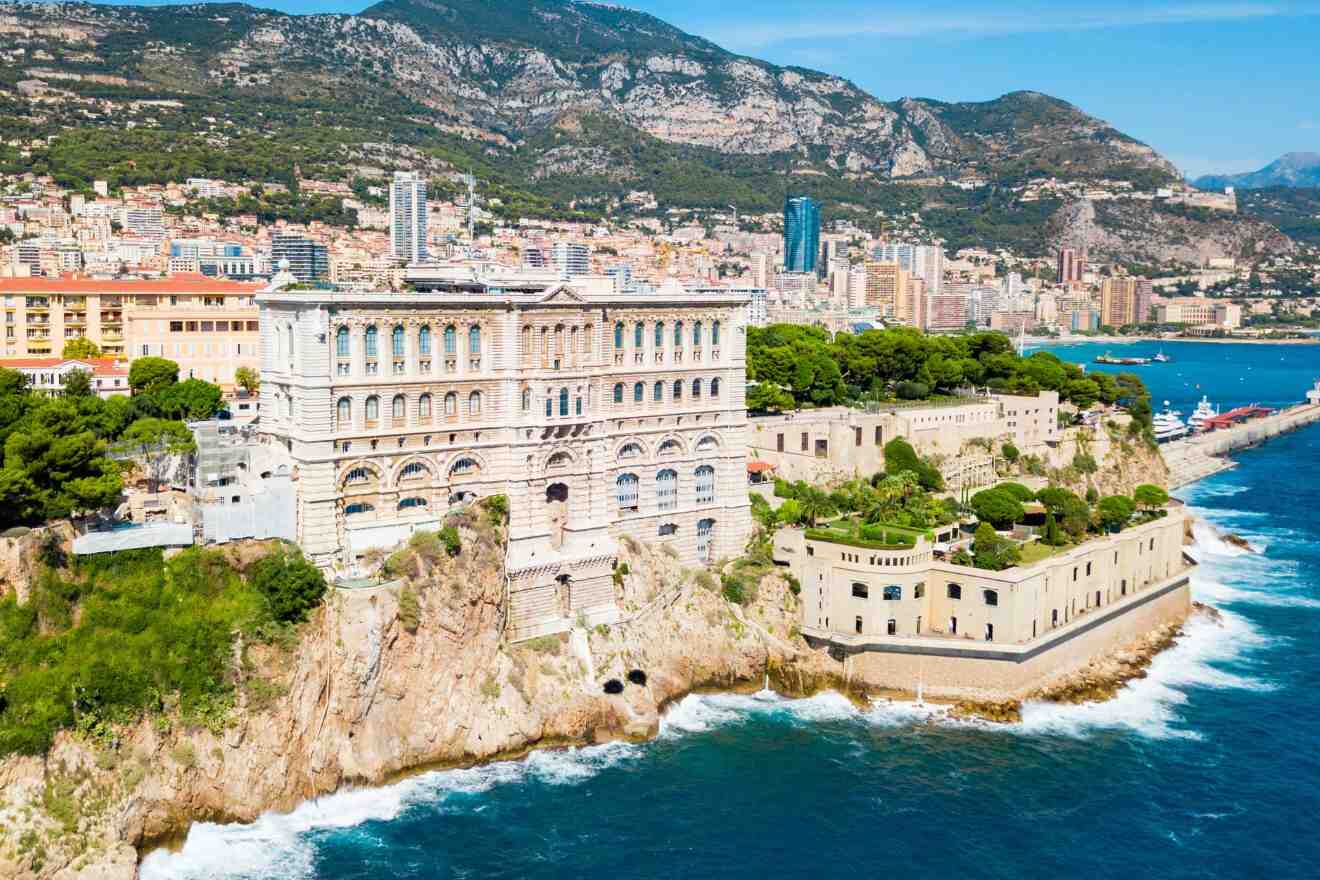 A grand, historical building perched on a cliff, overlooking the Mediterranean Sea with Monaco's cityscape and mountains in the background.