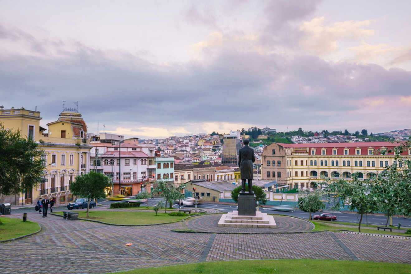 A cityscape features a central statue in a park with cobblestone paths, surrounded by multi-story buildings and rolling hills under a cloudy sky.