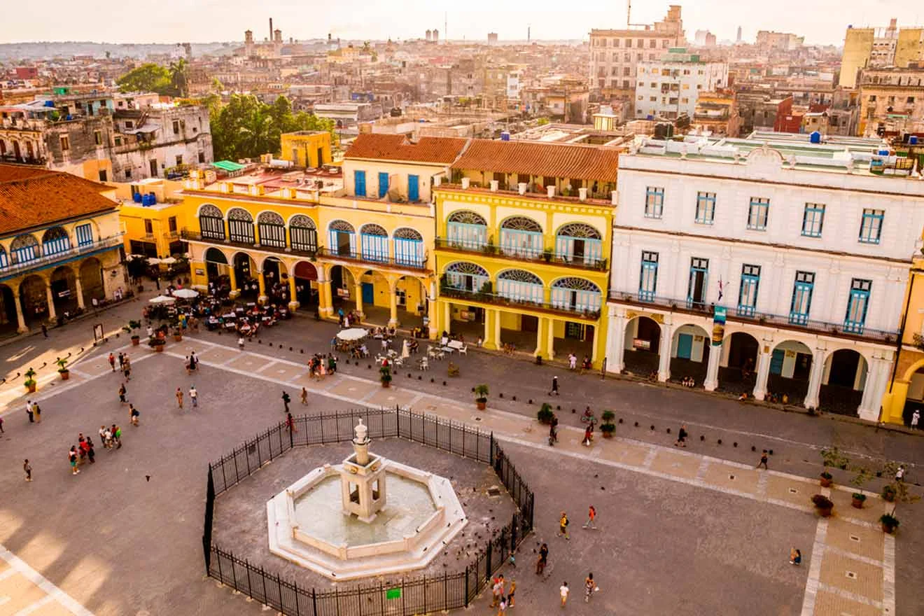 Aerial view of a colorful historic square with a central fountain surrounded by colonial-style buildings and people walking around.