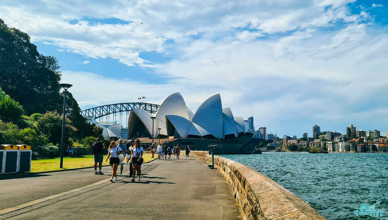 A pathway along the waterfront in Sydney with people walking and the Sydney Opera House and Harbour Bridge in the background on a sunny day.