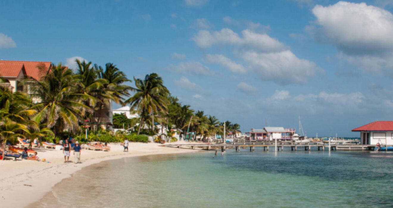 A sandy beach lined with palm trees, coastal buildings, and people. A pier extends into clear, calm water under a partly cloudy blue sky.